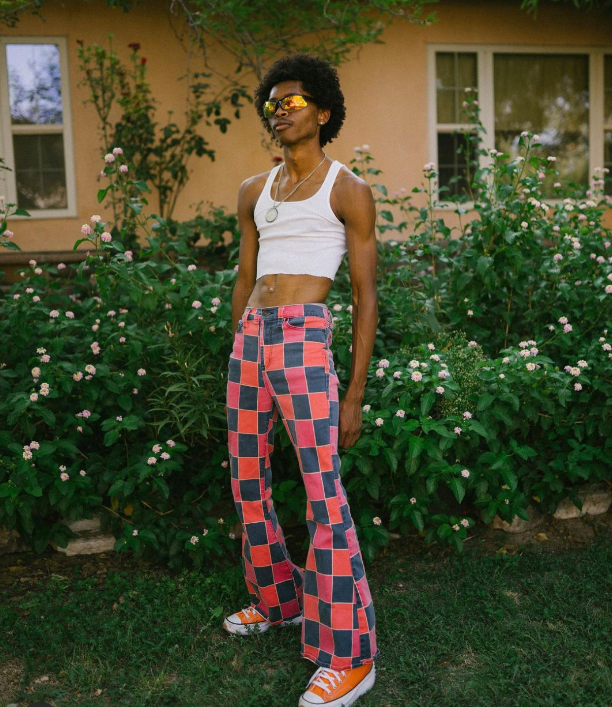 Katriel stands in front of a suburban home wearing a cropped white tank top, black and red checkered jeans, and orange Converse sneakers