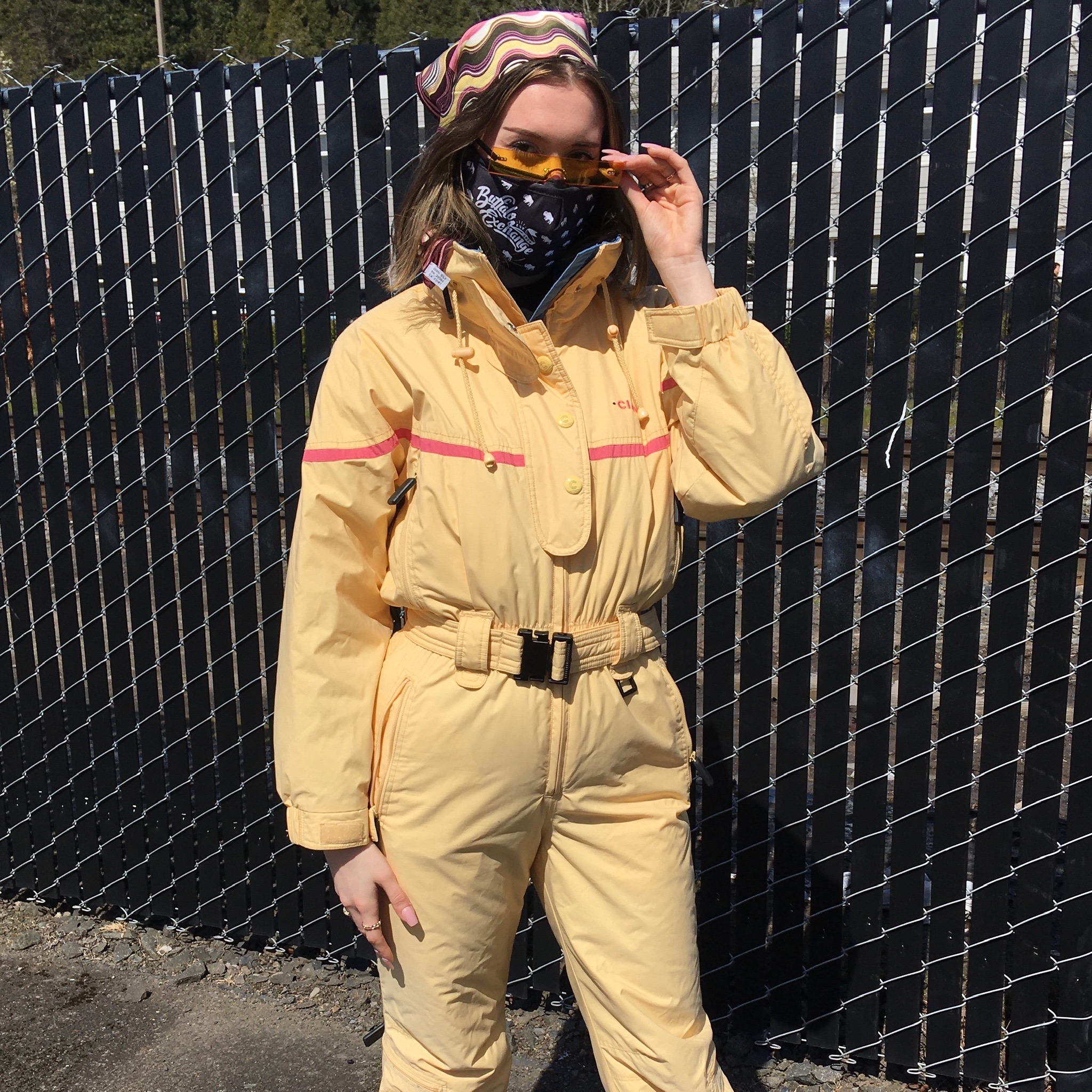 Person standing in front of wire fence wears a butter-yellow ski suit from the 80s and yellow-tinted rimless sunglasses