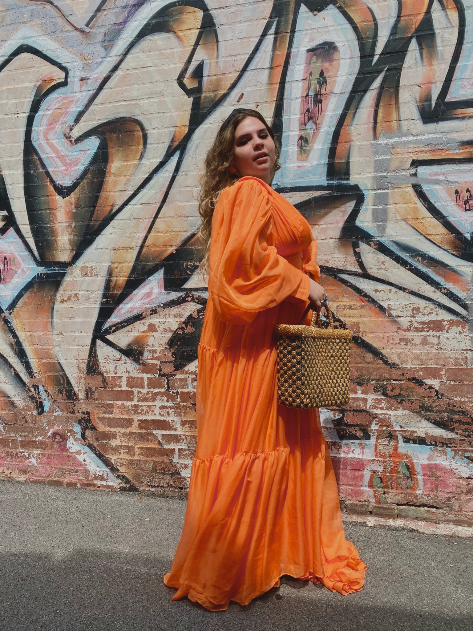 Person standing in front of graffiti wearing orange maxi dress and holding basket bag