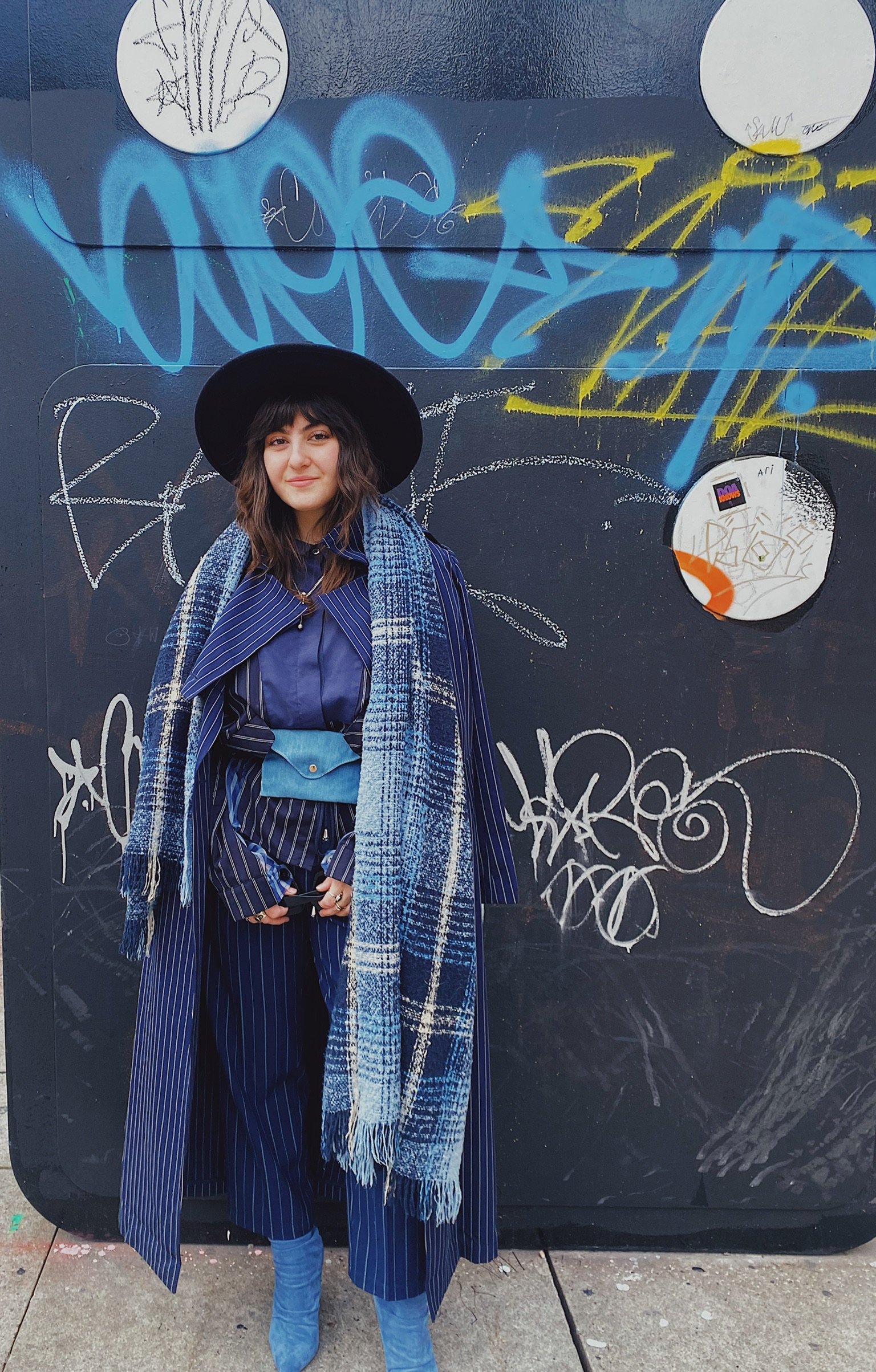 a person wearing a blue pinstriped suit layered with a blue plaid scarf and a black hat