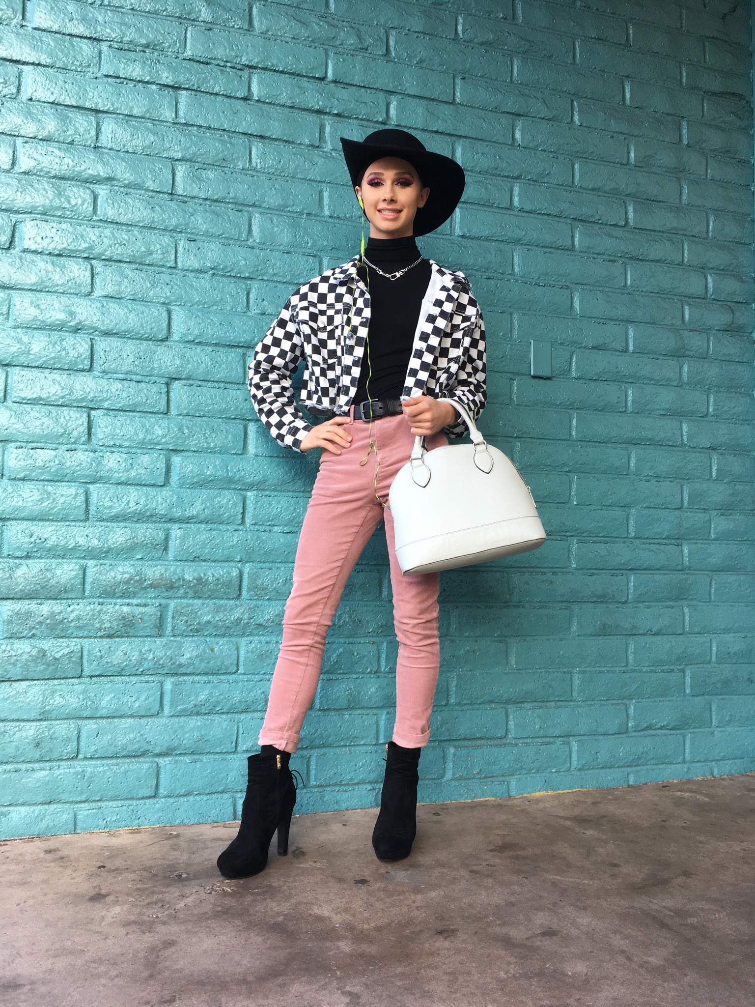 A person wearing black and white checkered jacket, pink pants, a black hat and a white handbag