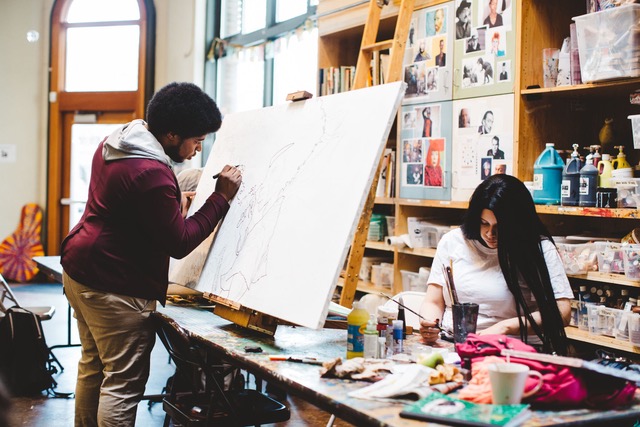 to the right a woman sitting at a table painting and to the left a man standing and paining on a blank canvas