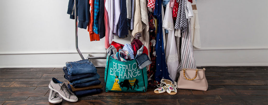 A clothing rack full of clothes and on the floor there are a pair of sneakers, a pile of jeans, a buffalo bag full of clothes, a pair of sandals and a handbag