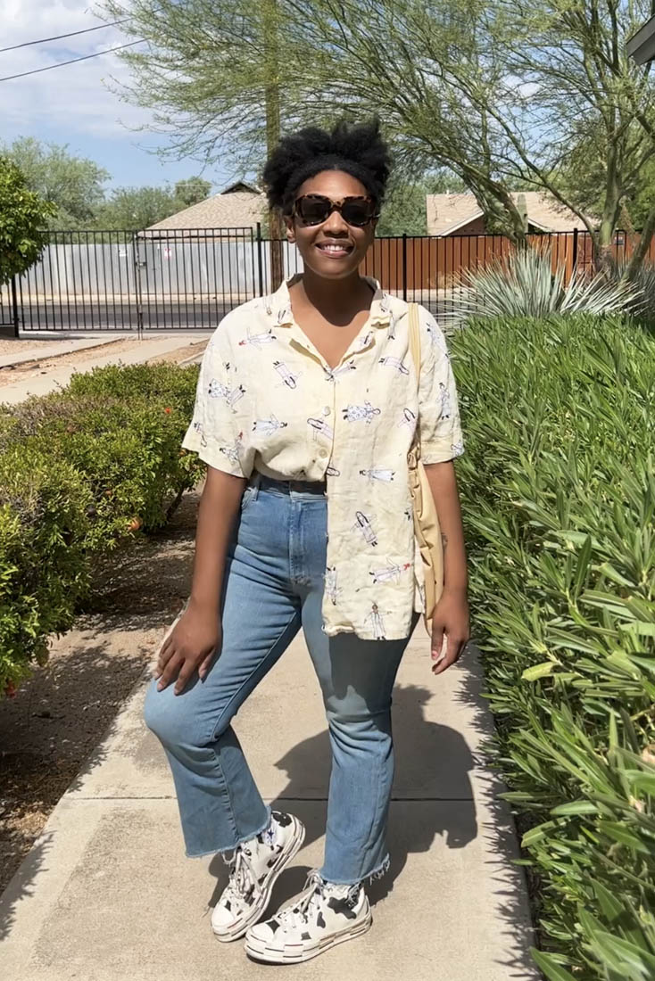 Person standing on sidewalk wearing crop flare jeans and half-tucked printed button-up with cow-print Converse sneakers