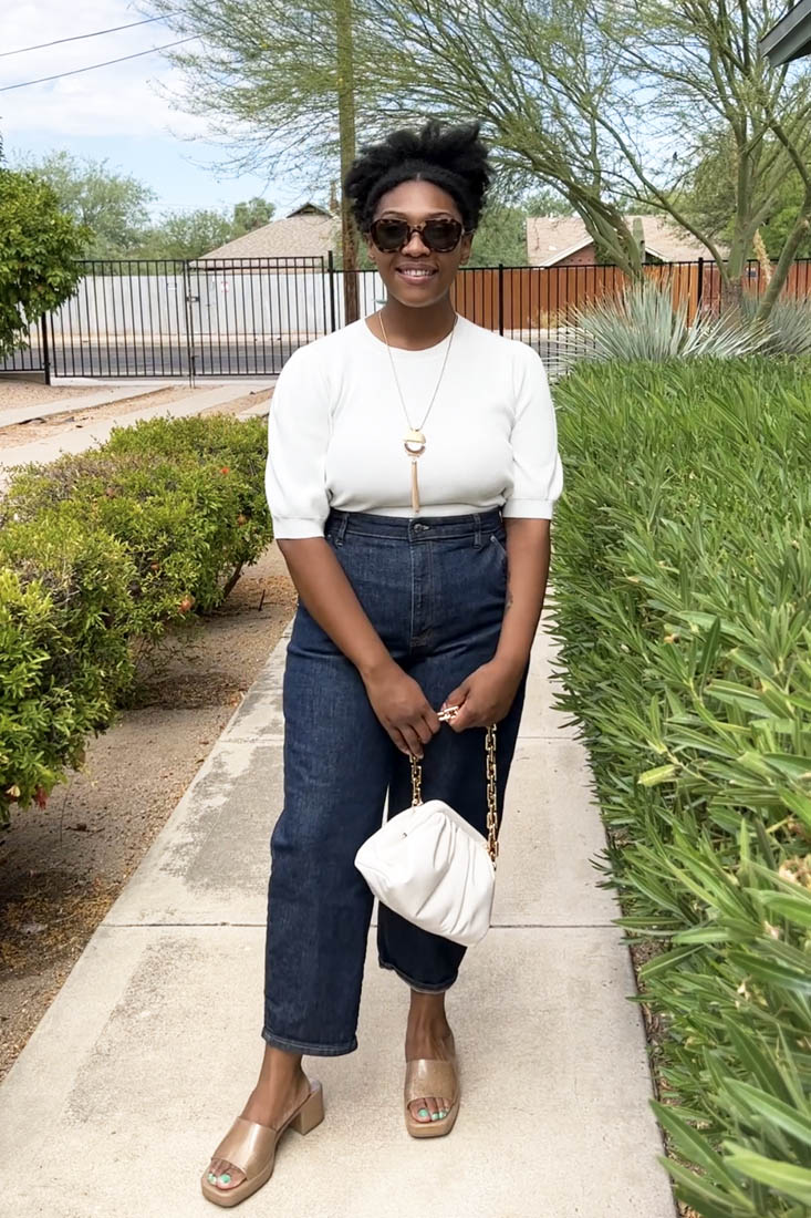 Person standing on sidewalk wearing white blouse with dark wash jeans, gold sandals and a white bag