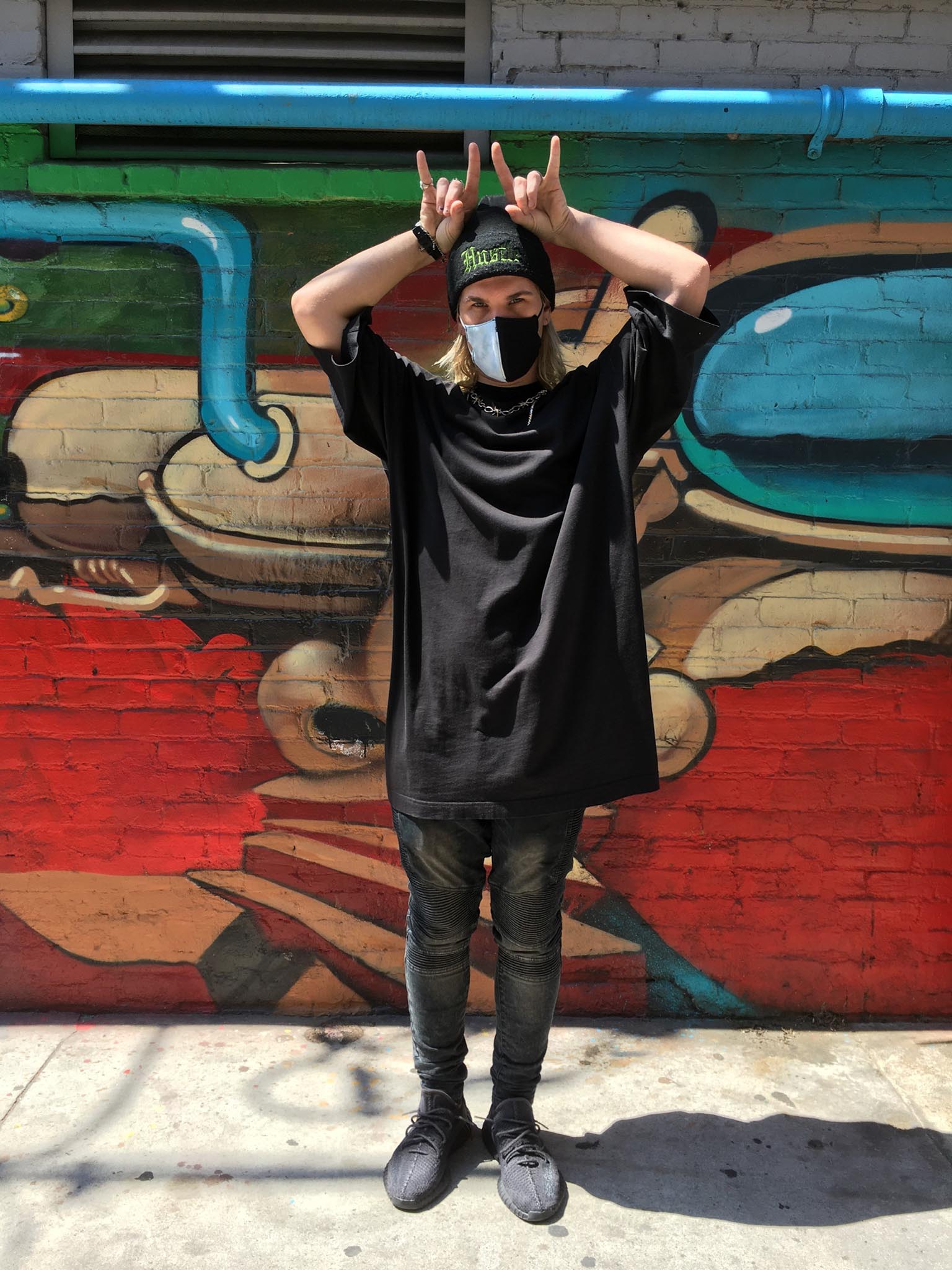 Person standing in front of mural wearing oversized black shirt, black beanie and black and white face mask