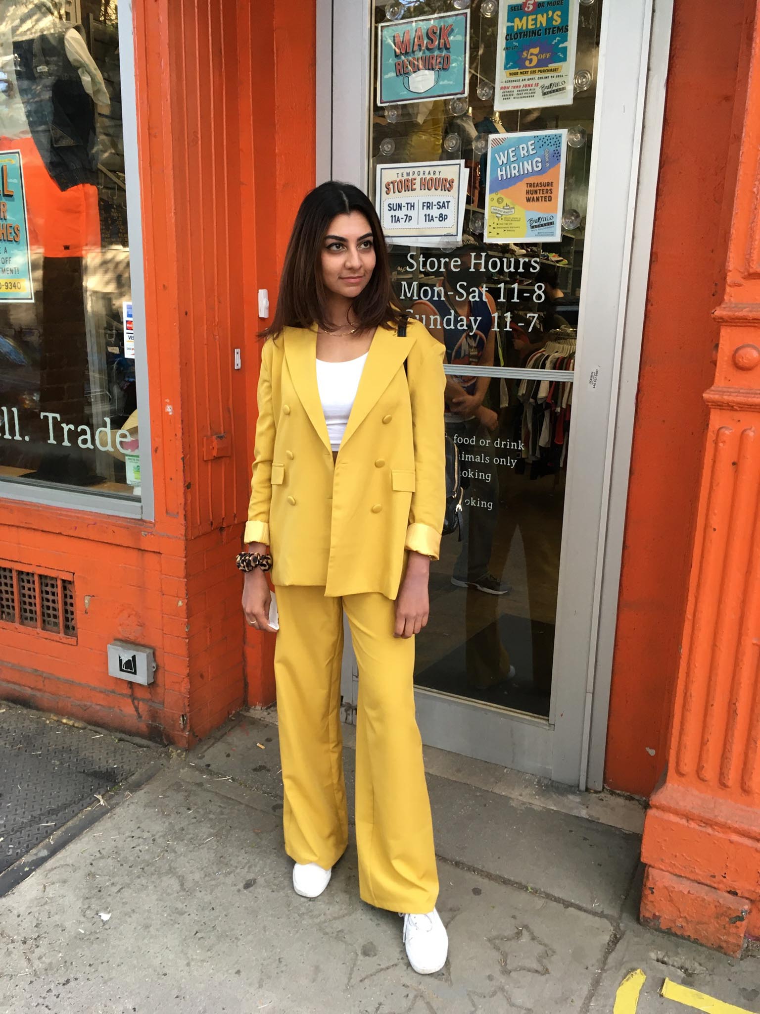 Person standing in front of orange shopfront wearing a mustard double-breasted suit with white crop top underneath and white sneakers