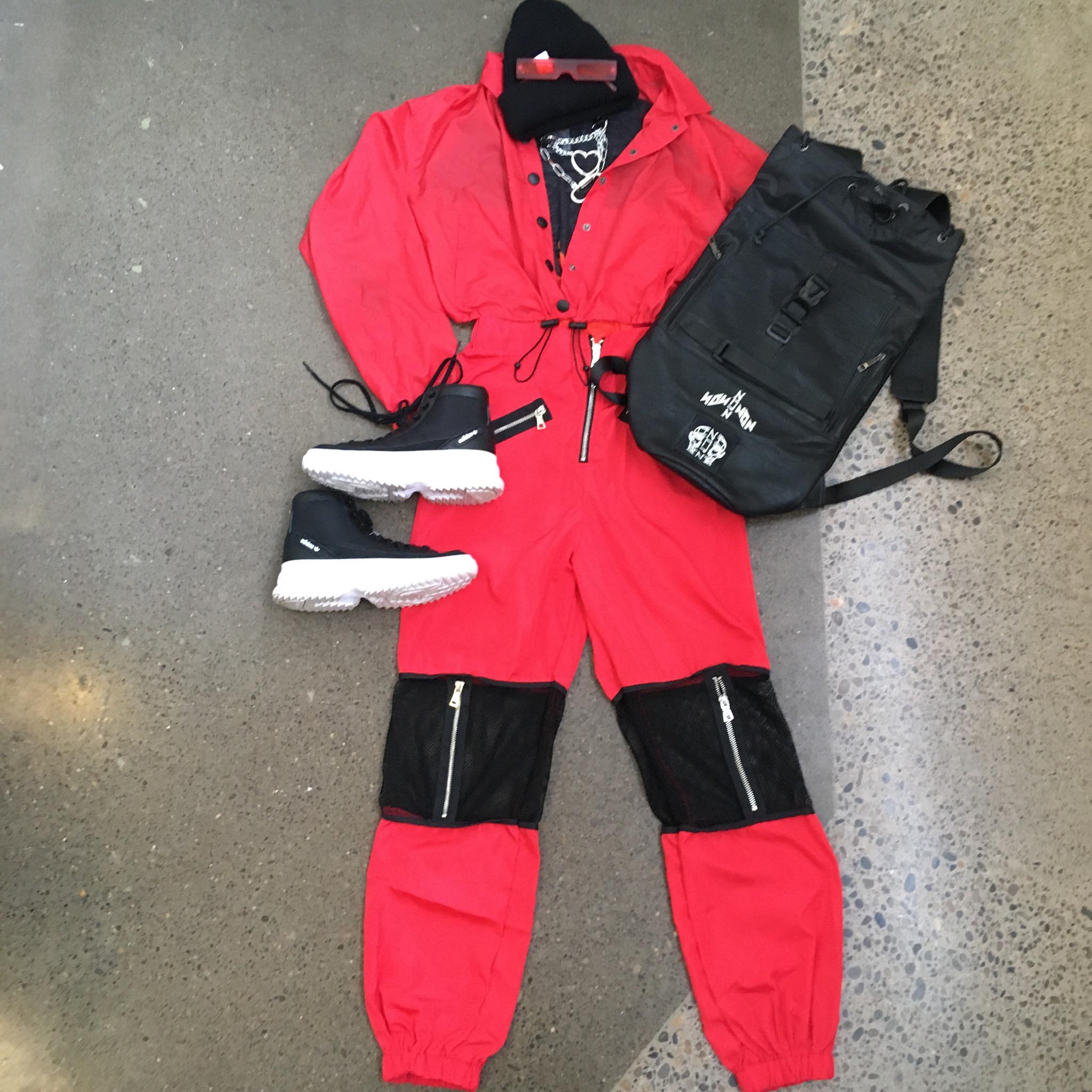 camera pointed down at bright red ski suit styled with white-sold hiking sneakers, silver chain necklaces, black backpack and beanie