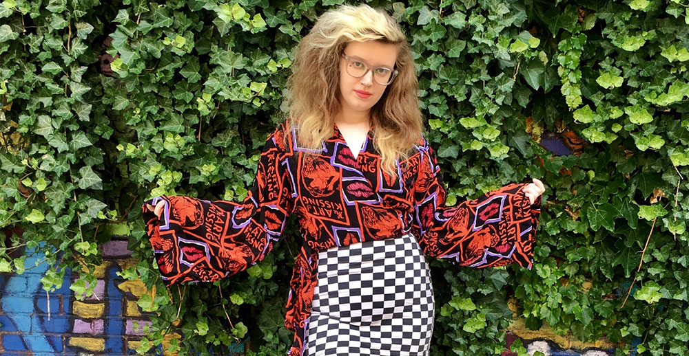 Brooke Posing in front of a green leaf wall, she is wearing a bold printed blouse and a black and white checkered skirt