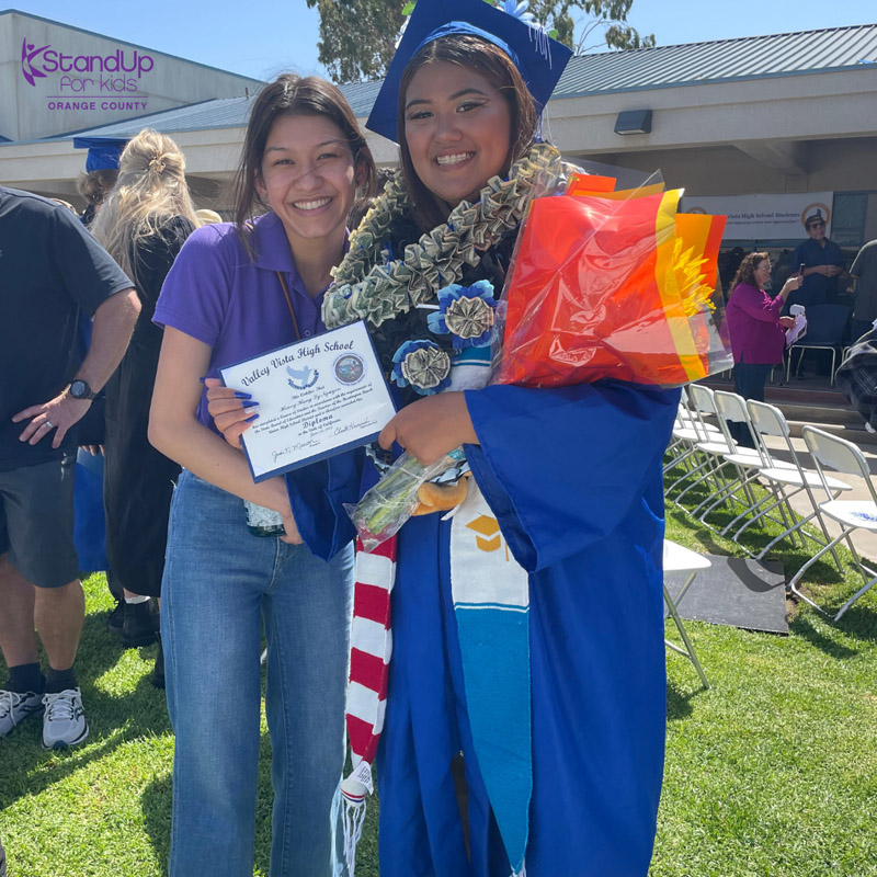 Two people at a graduation ceremony, one of them a StandUp for Kids graduate in cap and gown holding their diploma