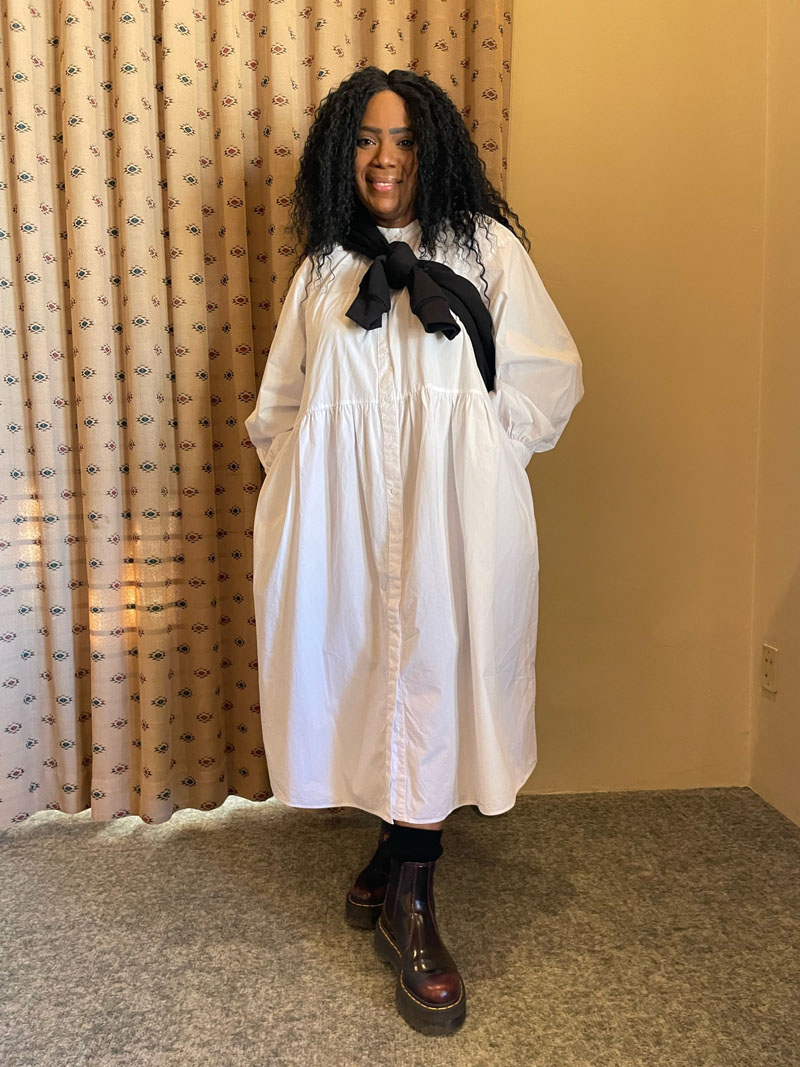 person standing against draped window wearing white cotton midi dress, burgundy lace up boots and black sweater tied around their neck