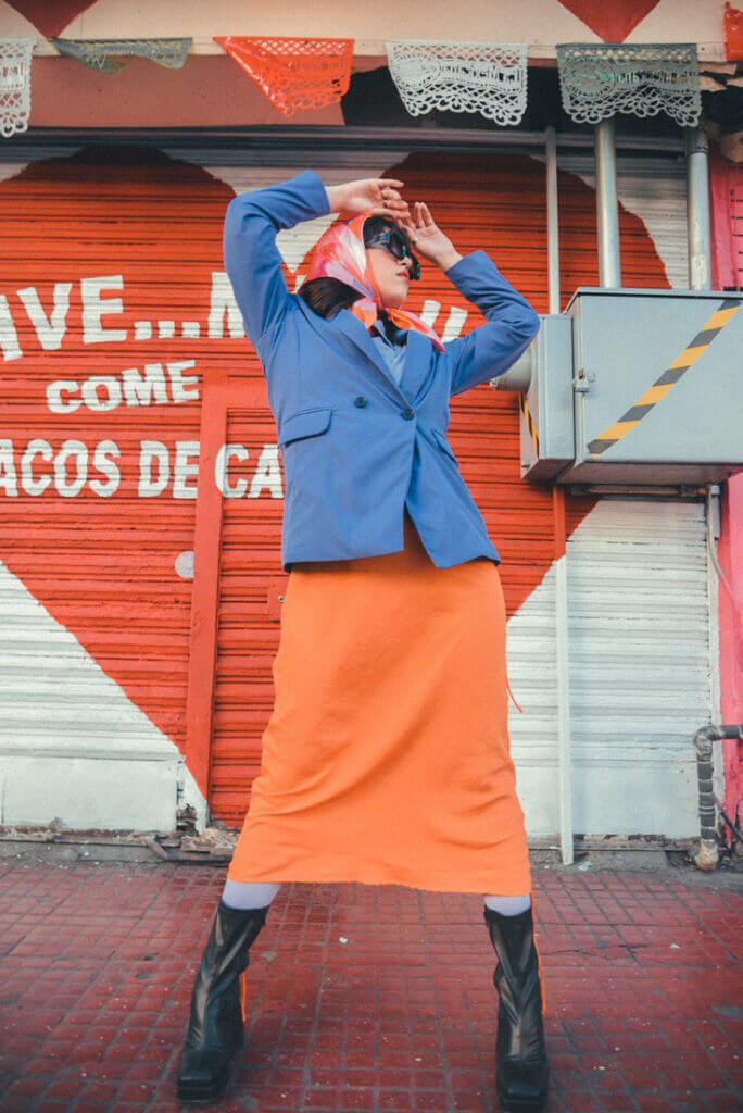 Person with their hands above their head wearing a red plaid scarf tied around their head, large sunglasses, a blue blazer buttoned up, and a long bright red skirt.