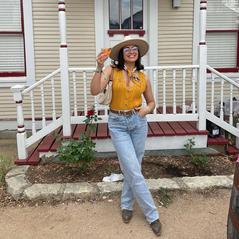 @cozyrosati wearing a yellow cable-knit sweater vest, light wash straight leg jeans, a yellow handkerchief, and a wide brim hat.