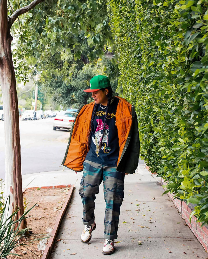 Customer @jerrywiththegoodhair wearing a quilted black and copper jacket, Ed Hardy graphic tee, camo cargo pants, and white Doc Martens