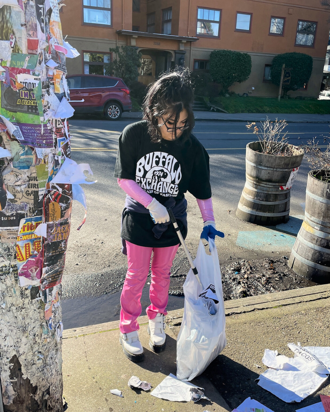 Buffalo Exchange employee in a Buffalo Exchange tee and pink pants picking trash off the sidewalk with a trash grabber