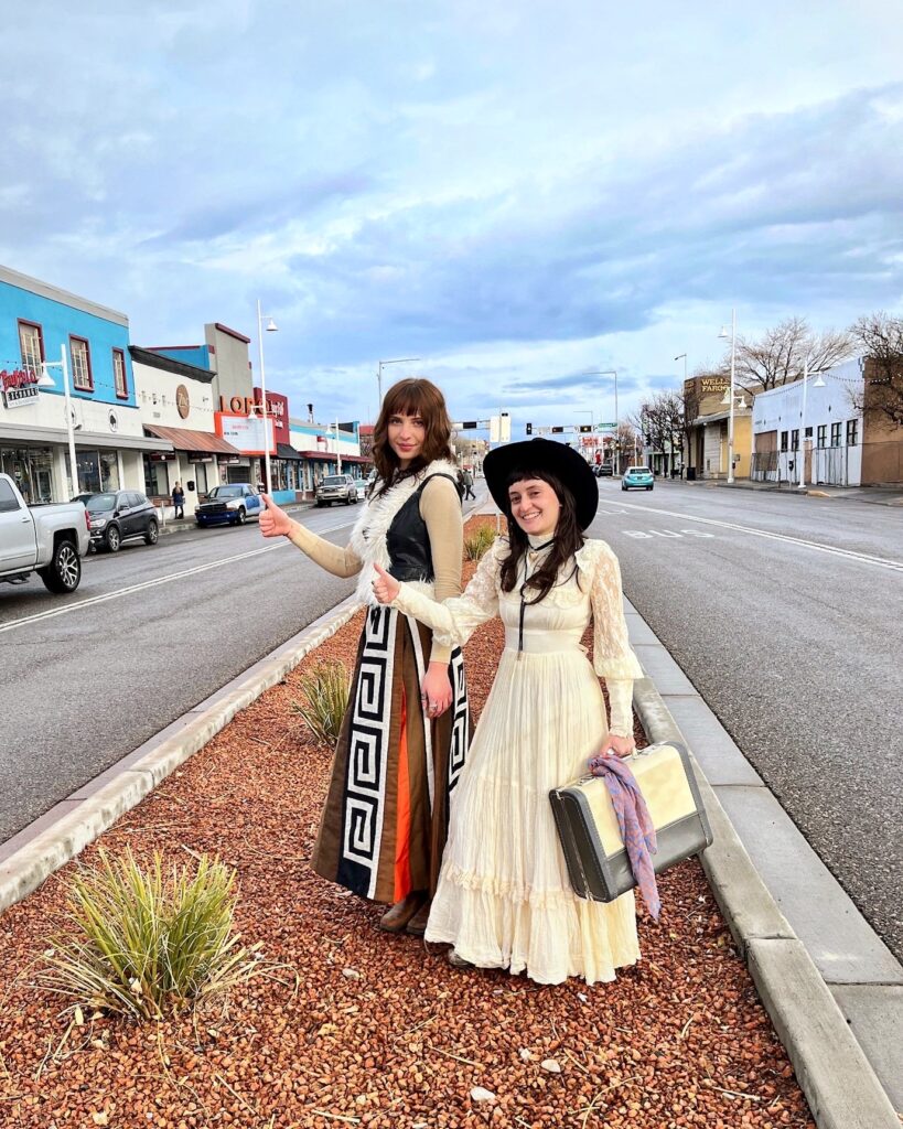 Two women dressed in 70s outfits hitchhike on the street