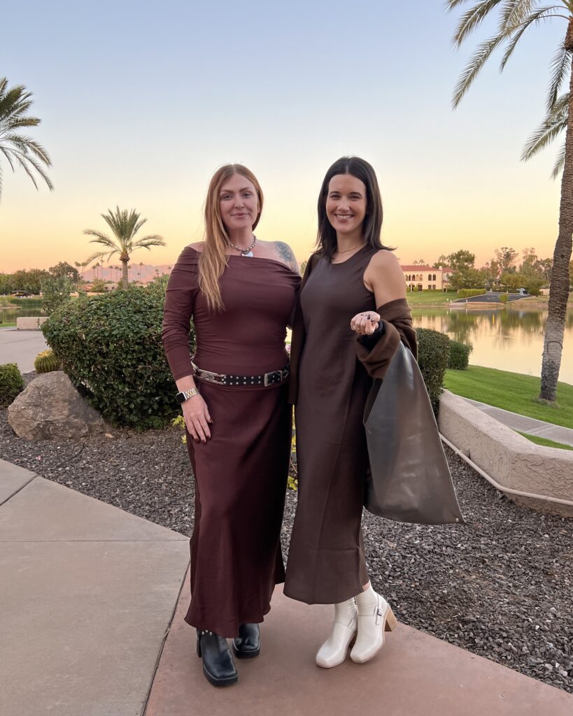 Two women wearing brown semi formal outfits pose in front of the sunset