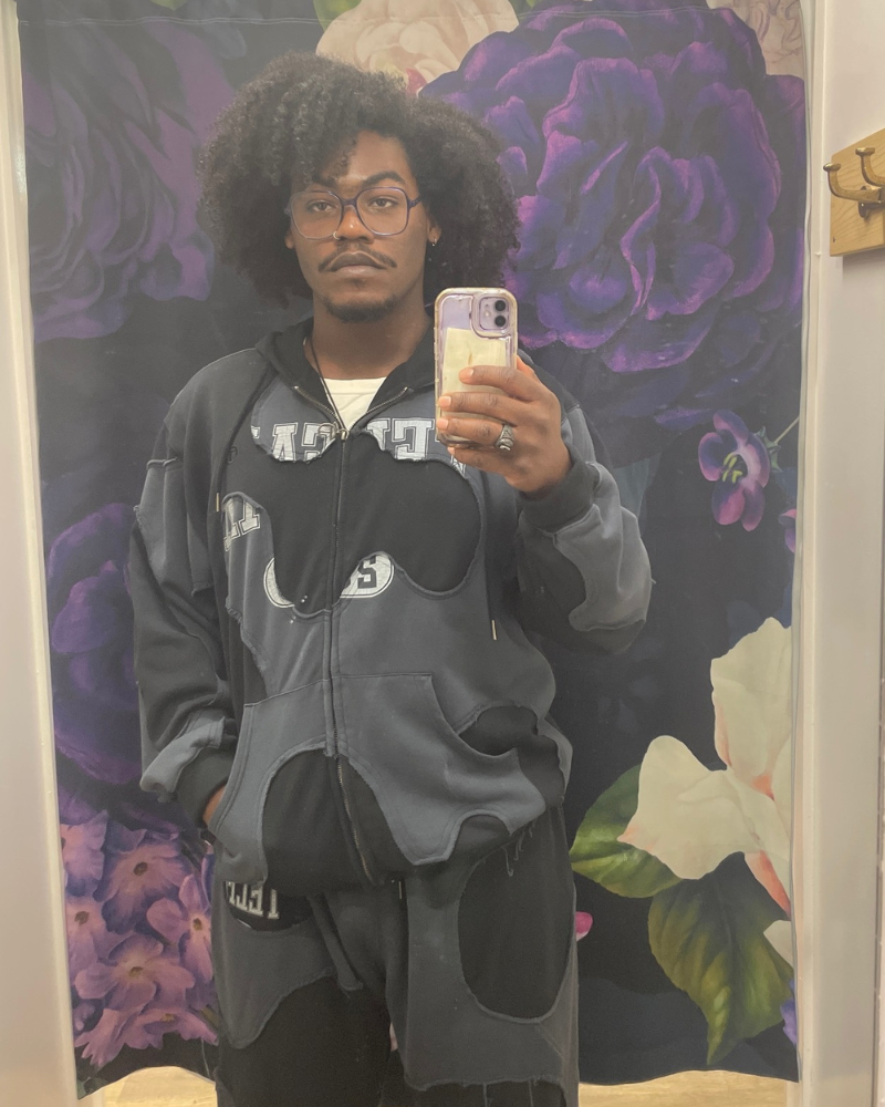Untitled design (2) Person in a fitting room with floral curtain takes a mirror selfie with iPhone
