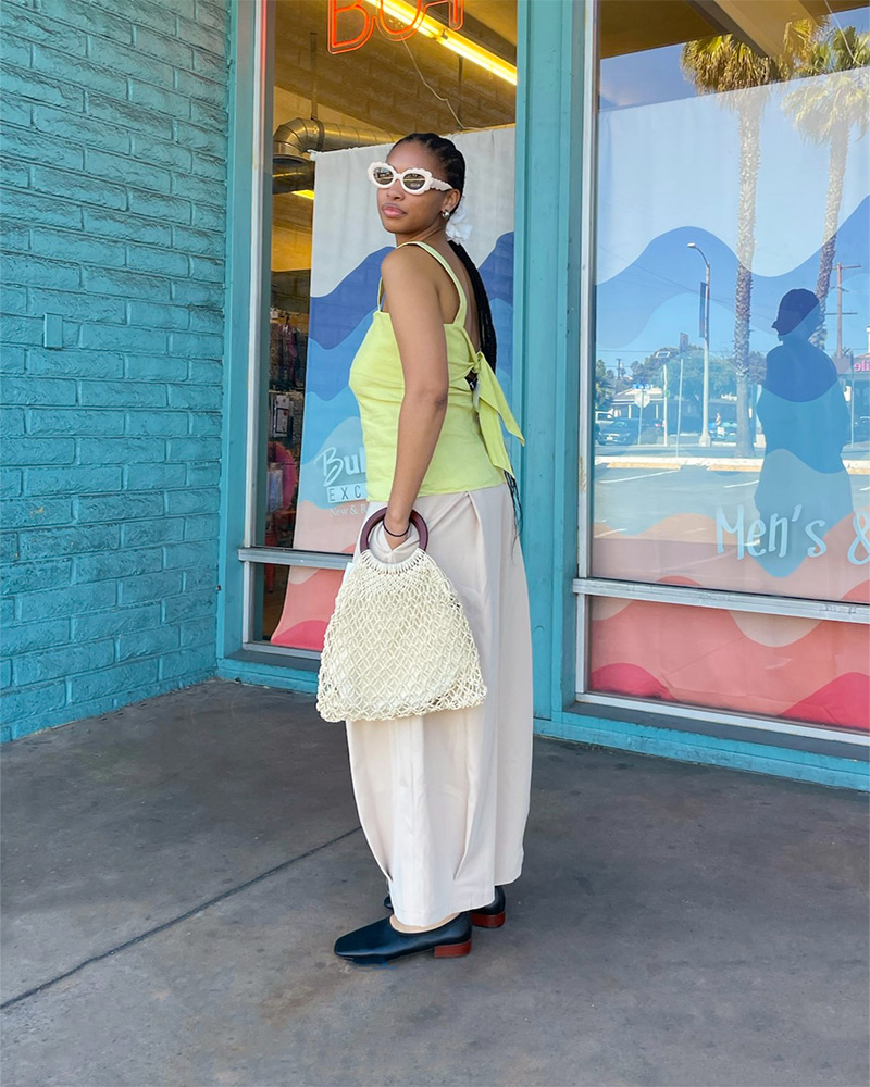 Blog-15-Edited A woman wears a light green sleeveless top, beige pants, a woven tote bag and black flat shoes