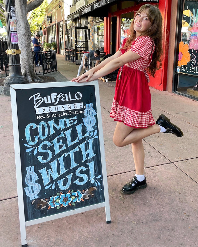 Blog-27-Edited A woman poses with a sidewalk sign that reads "Buffalo Exchange Come Sell With Us $"