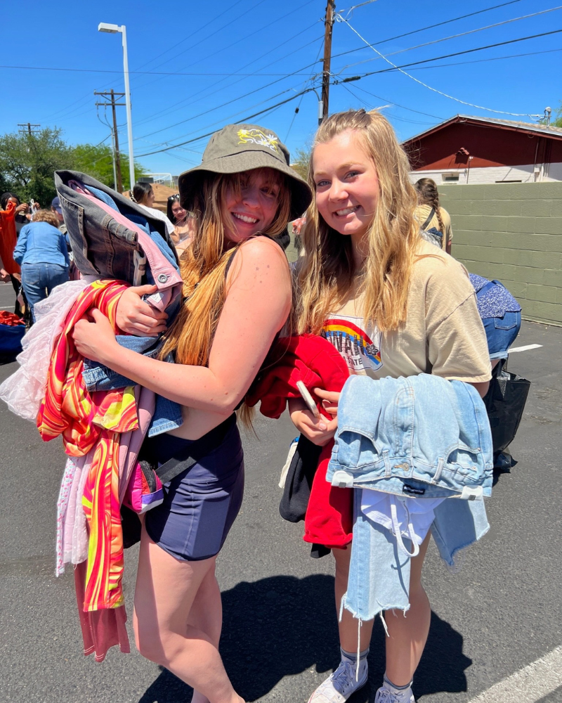 eday blog pic 3 resized two friends smile at camera while holding clothing in a parking lot