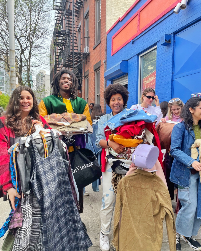 eday blog pic 5 resized three friends smile while holding armfulls of clothing near a blue brick building