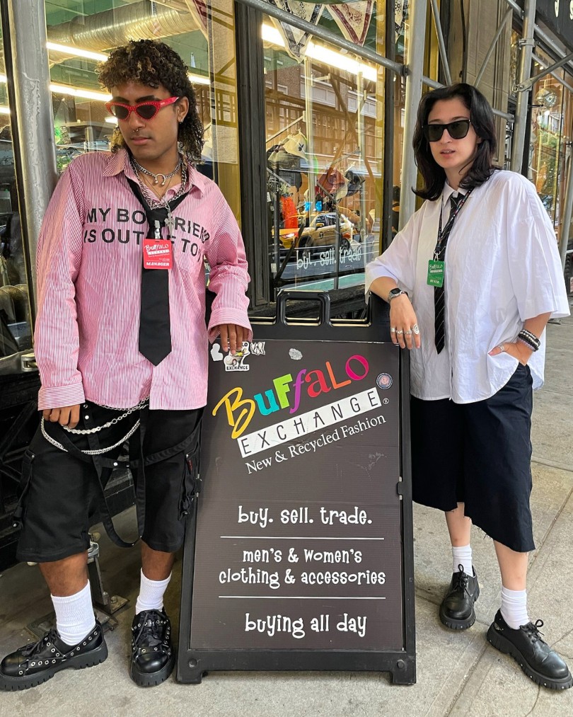 Two employees in front of Buffalo Exchange wearing wrinkled oversized button downs, loose ties, silver chains, black knee-length shorts and loafers with ankle socks