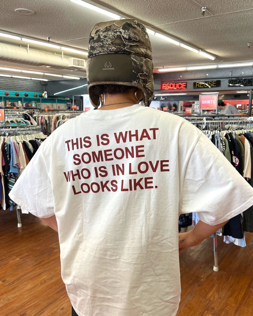 A person inside Buffalo Exchange showing the back of their graphic tee shirt that says in capitalized red letters: "This is what someone who is in love looks like."