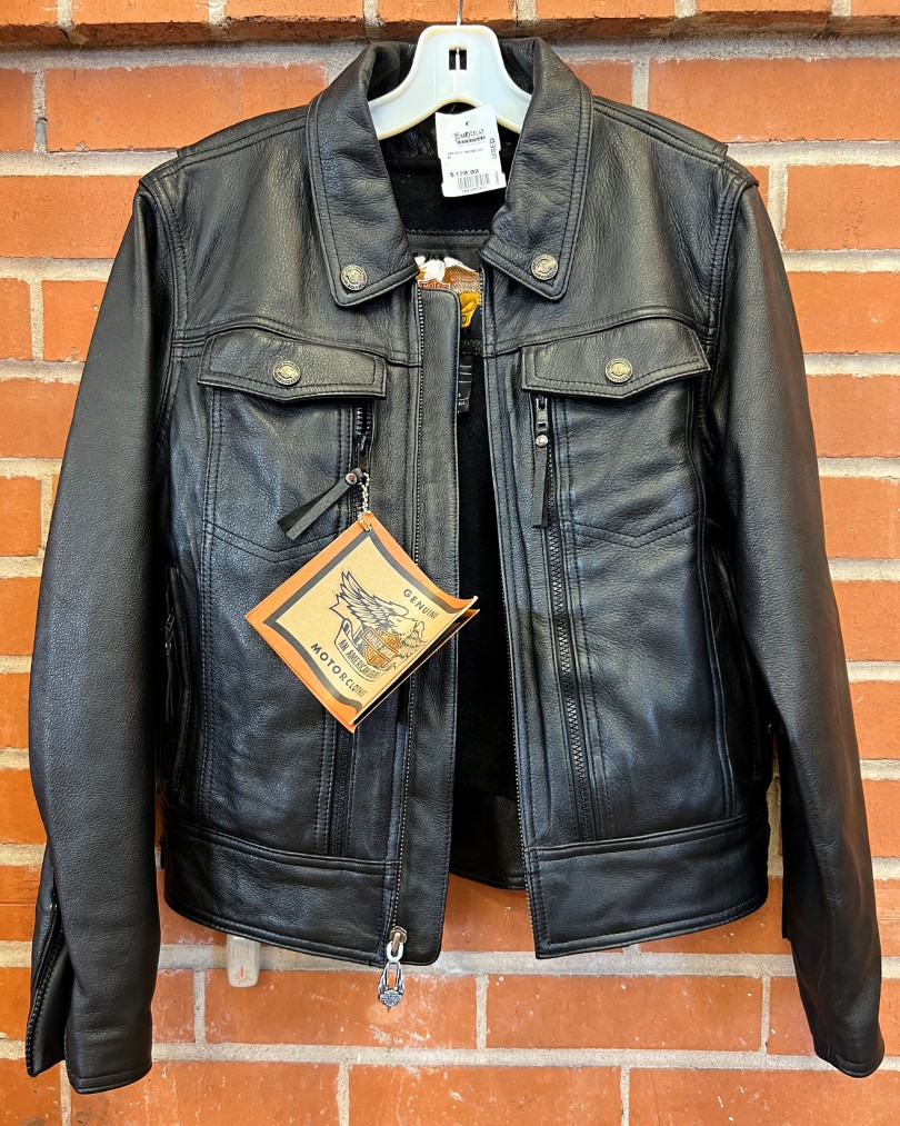 A brand new black leather Harley Davidson motorcycle jacket with tags attached hanging against a brick wall at Buffalo Exchange
