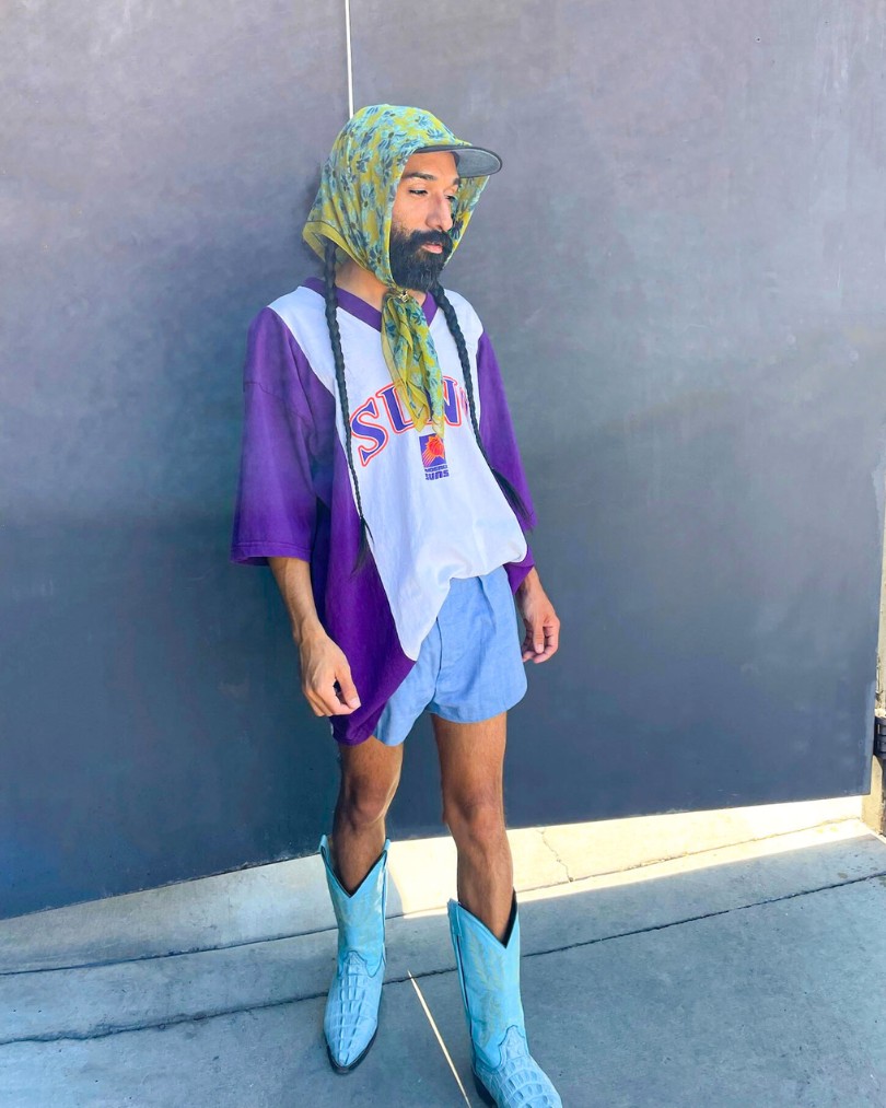 Person wearing an oversized Phoenix Suns tee shirt half tucked into blue shorts, turquoise western boots, and a baseball cap secured with a green floral scarf tied around their head