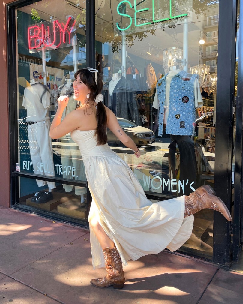 Person in front of neon signs that say "Buy" and "Sell" pretending to run into a Buffalo Exchange while wearing a strapless cream dress and brown leather western boots