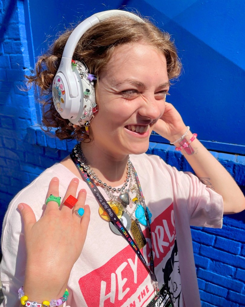 Person with maximalist style wearing a Betty Boop tee, over-the-ear headphones embellished with stickers, charm necklaces, beaded bracelets, and colorful rings