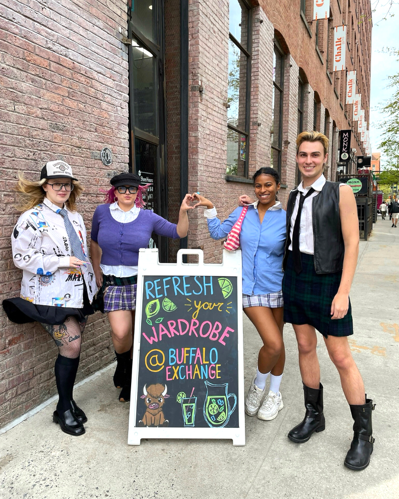 A group of Buffalo Exchange employees pose next to a sidewalk sign that reads "Refresh your wardrobe at Buffalo Exchange"