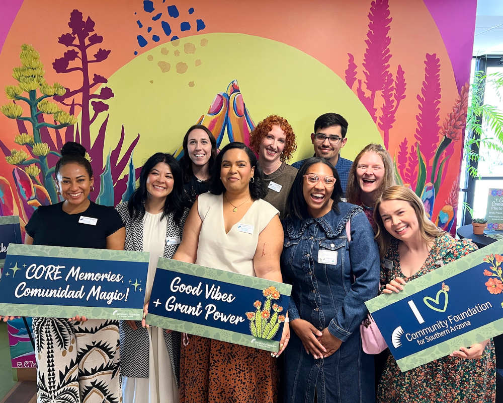 A group of people standing in front of a sunset mural smile and hold signs that read CORE Memories Communidad Magic, Good Vibes + Grant Power and I Heart Community Foundation of Southern Arizona